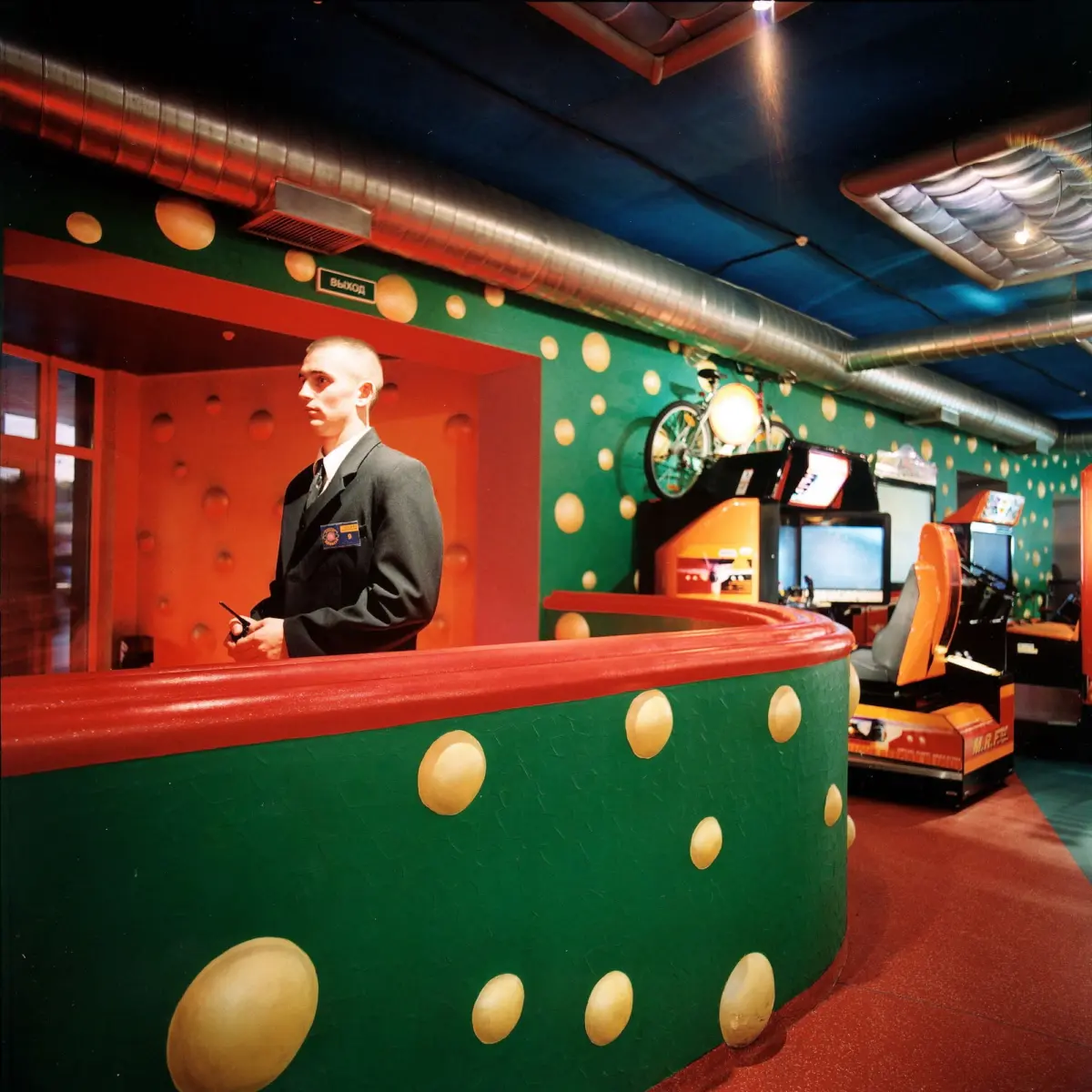 Bald mannequin in a suit stands behind a curved red-and-green counter in a retro arcade with arcade cabinets.