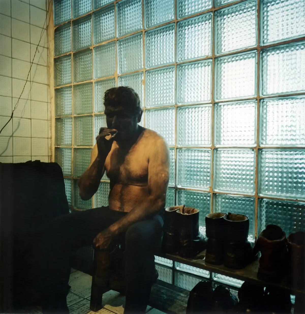 Shirtless man seated before a glass-block wall, touching his mouth with a hand; row of black leather boots on a shelf in the foreground.
