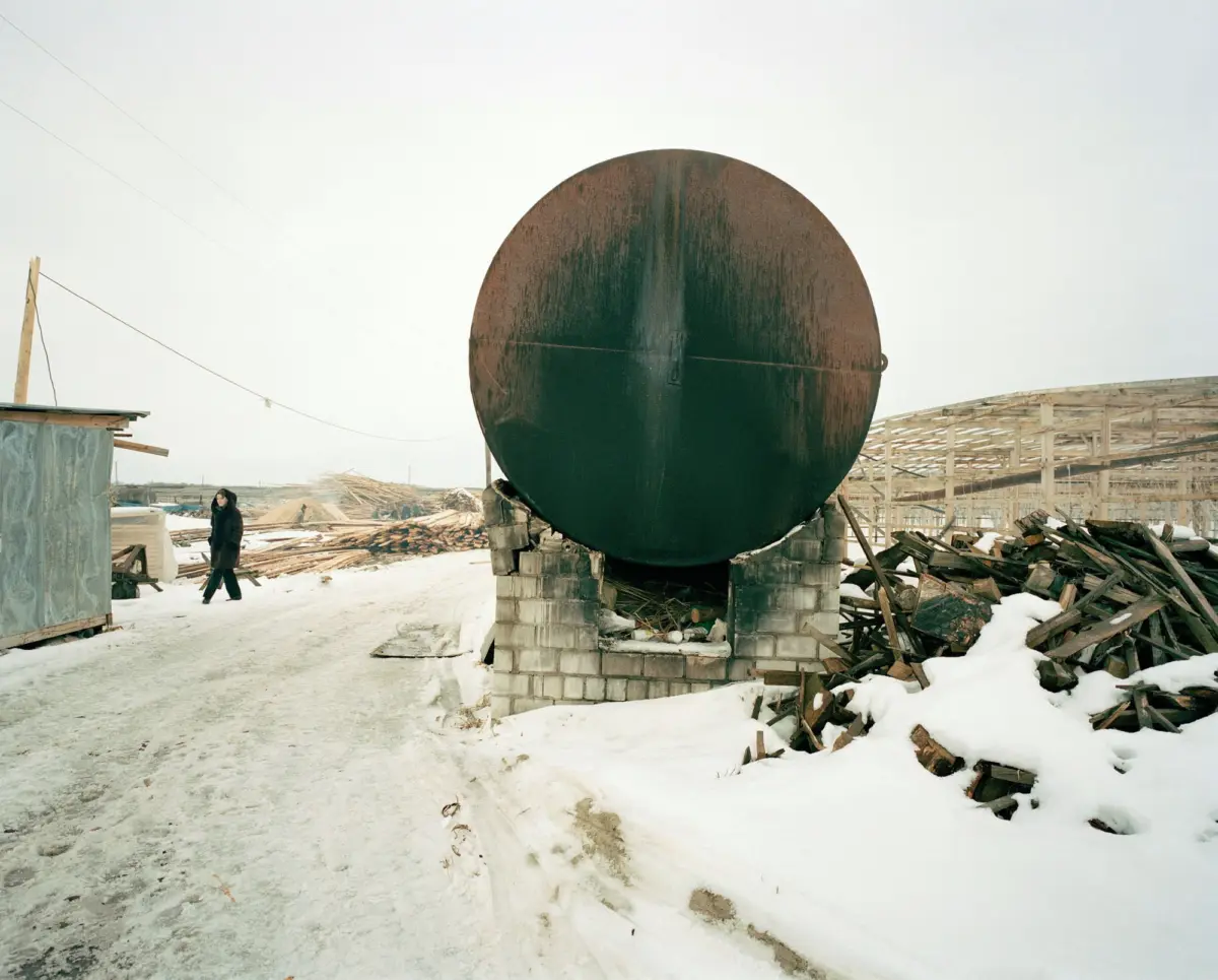 Large rusted circular steel storage tank on concrete supports at a snowy construction site; a person in a dark coat walks nearby.