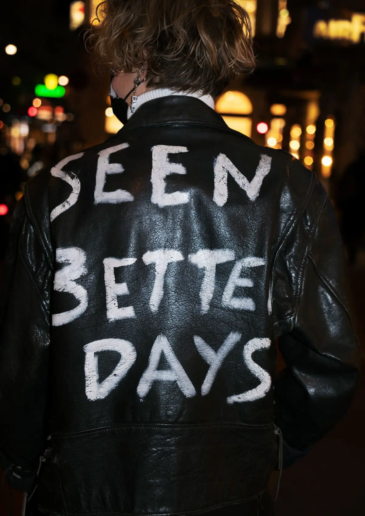 Person in a black leather jacket with "SEEN BETTER DAYS" painted on the back, walking away on a nighttime city street.