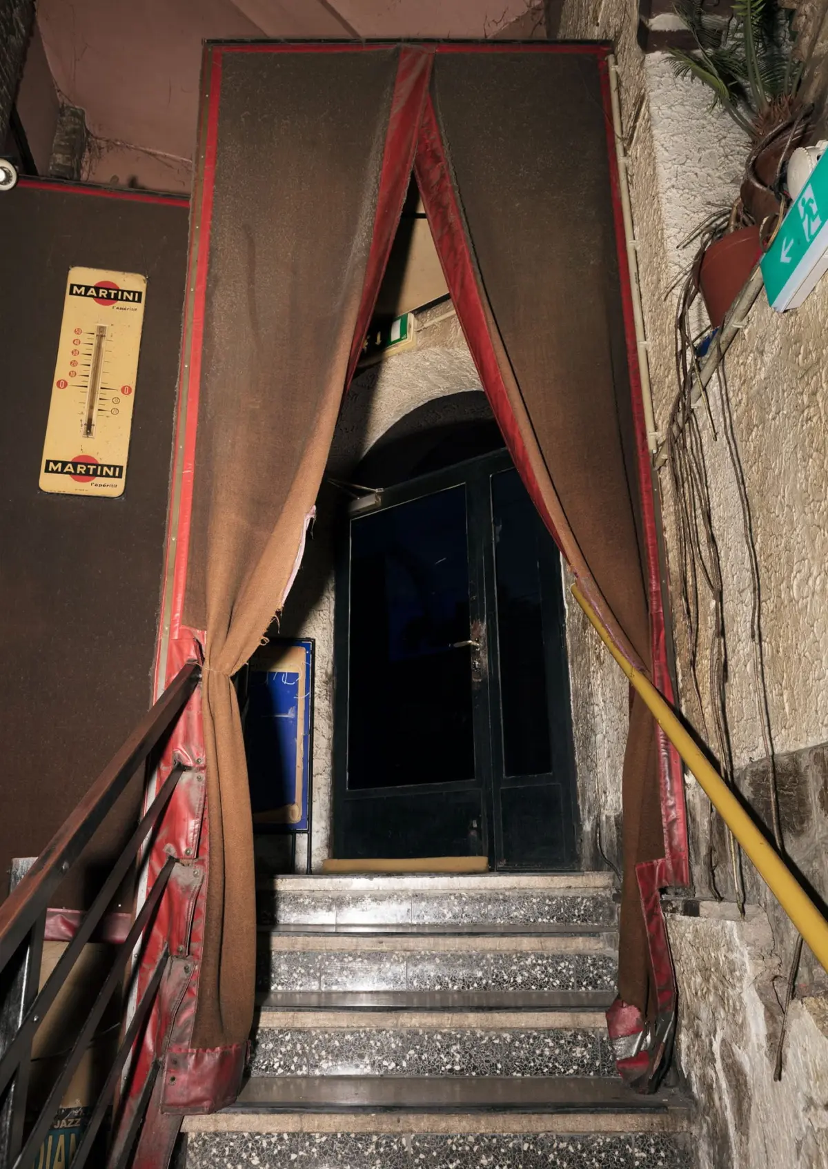 Dim stairwell entrance framed by red-draped doorway, terrazzo steps lead to a dark glass door; wall-mounted Martini thermometer sign on the left.