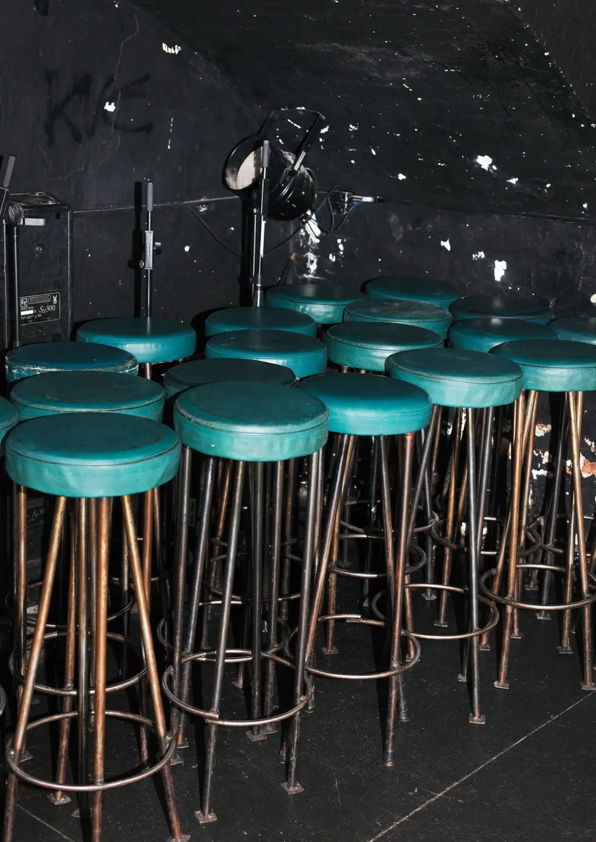 Row of teal round stools with metal legs on a dark backstage stage; microphone stand and audio gear in the background.
