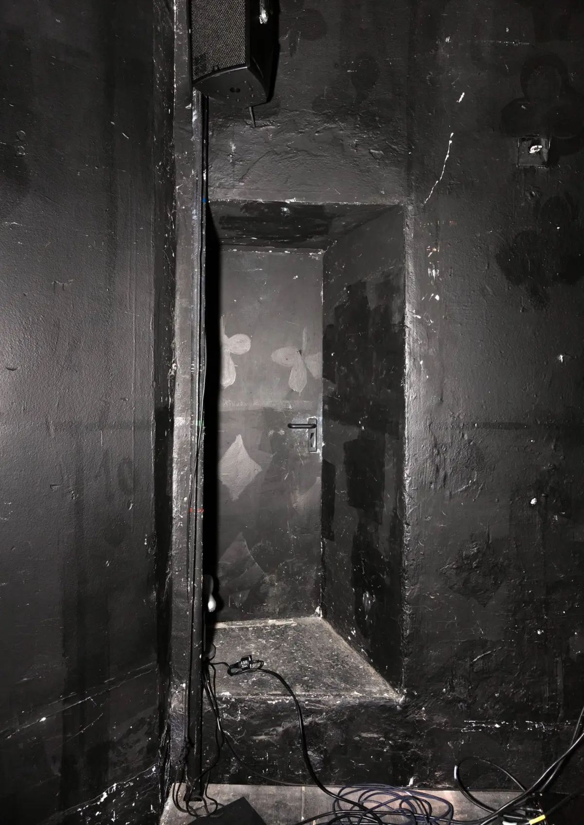 Dark backstage area with a wall-mounted speaker, a recessed doorway in a narrow stair alcove, and tangled cables on the floor.