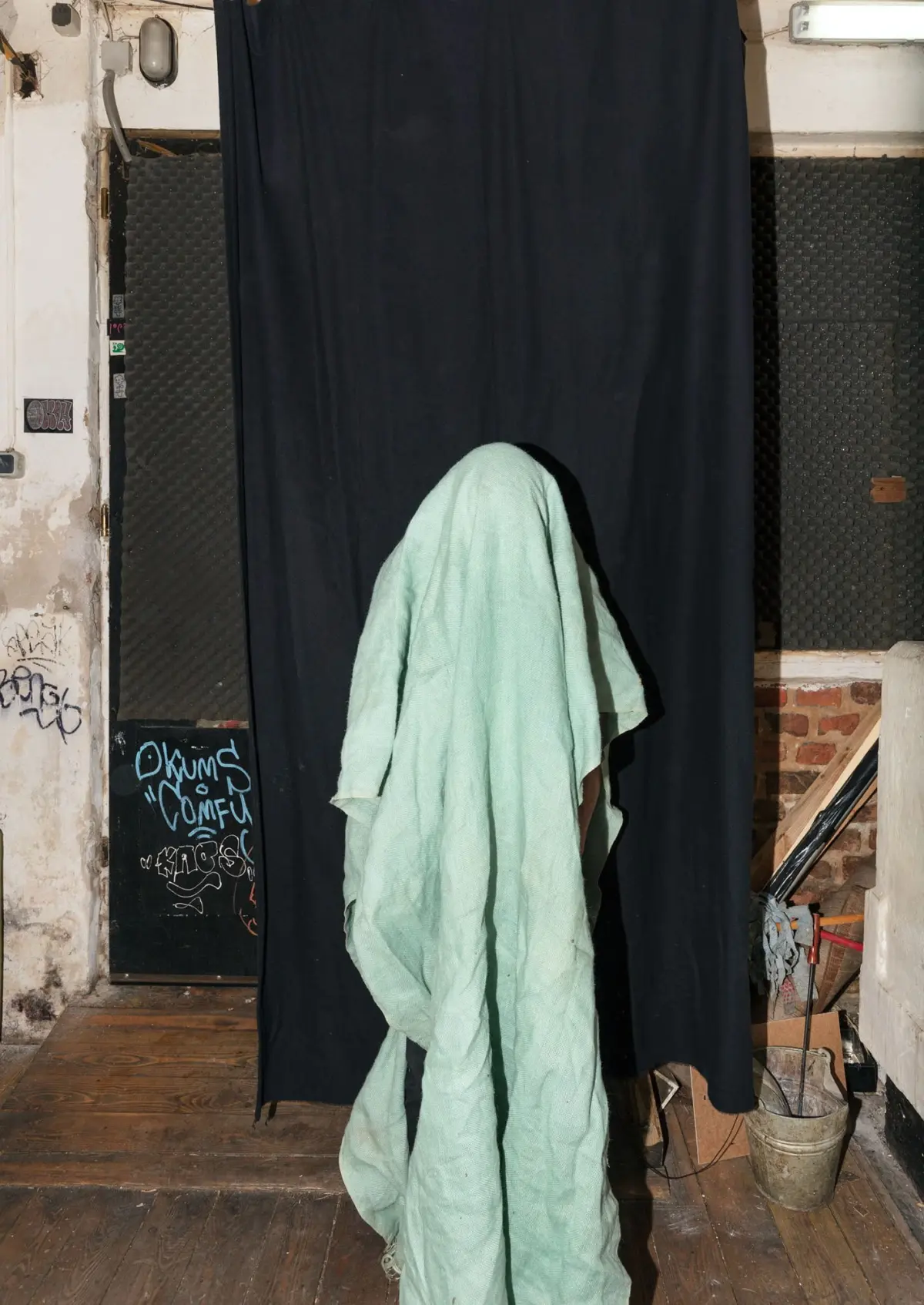 Figure draped in mint-green sheet stands before a black curtain in a gritty industrial interior with graffiti and a worn wooden floor.