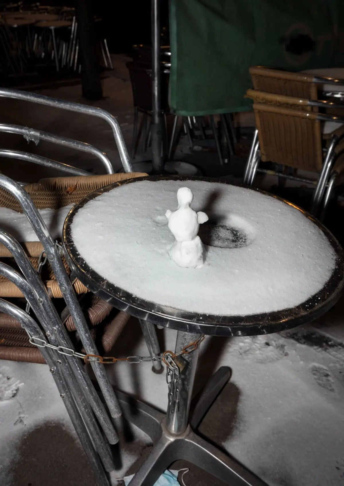 Snowman on a snow-covered round cafe table in an outdoor seating area at night.