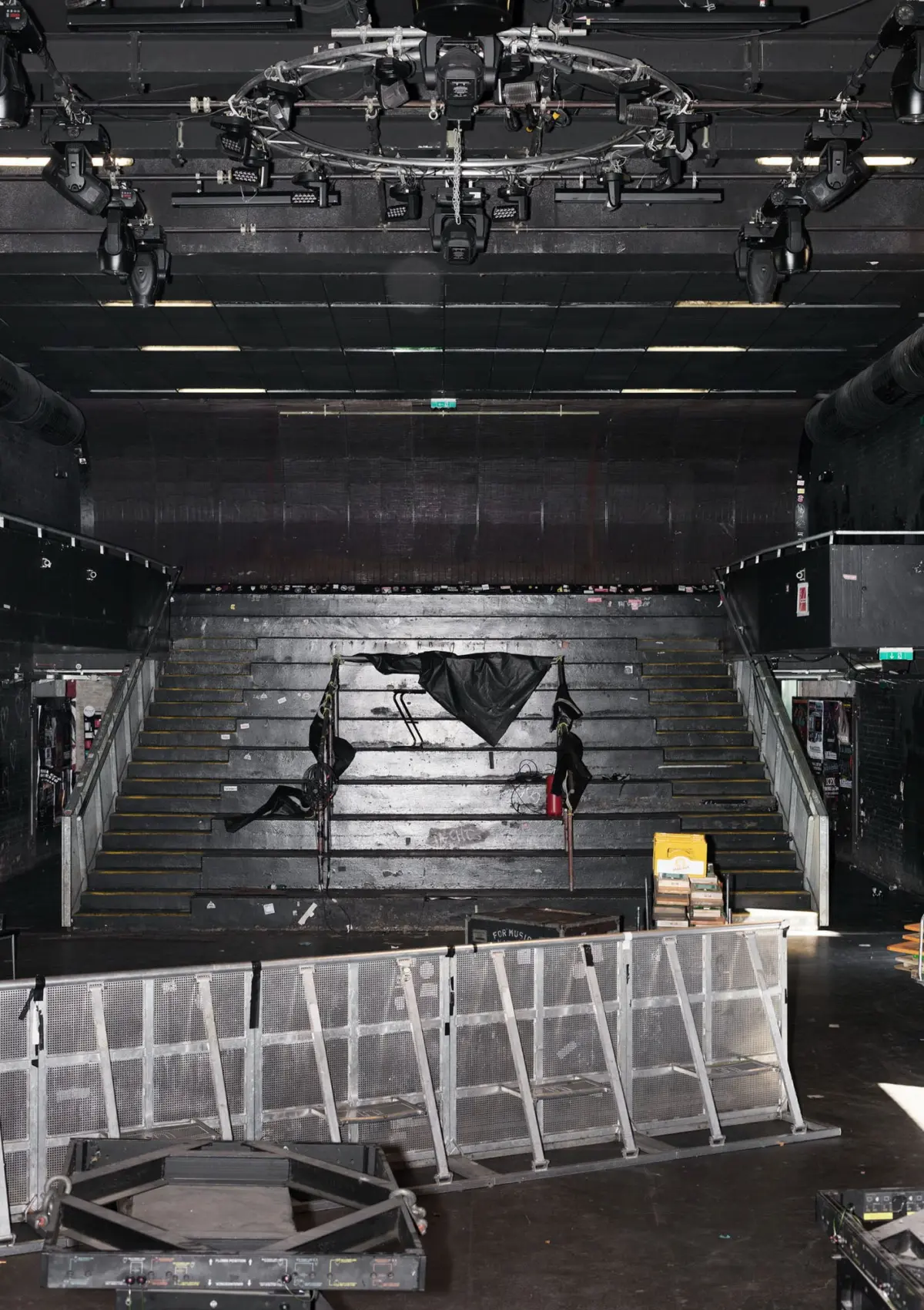 Backstage theater with overhead lighting rig and cables, empty dark stage, flanking stairs, and metal barricades; scattered gear.