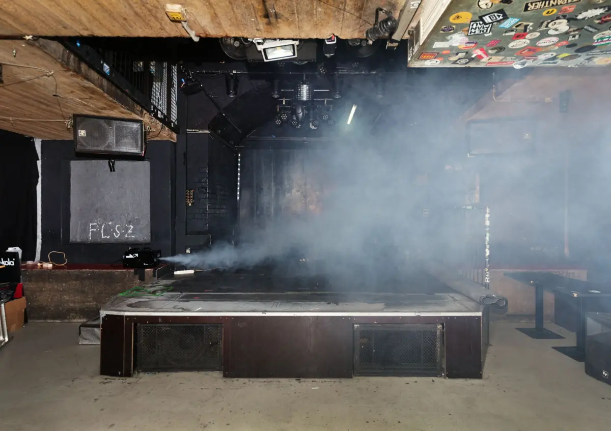 Stage with large black PA speakers and lighting rig, hazy with smoke in an indoor music venue.