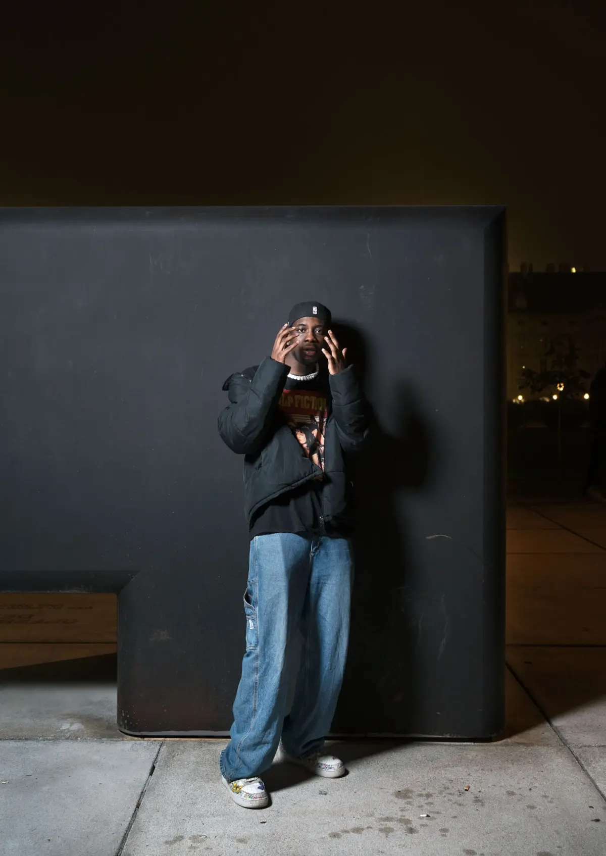 Person wearing a black jacket, cap, and baggy jeans stands against a large black wall at night, talking on a cellphone.