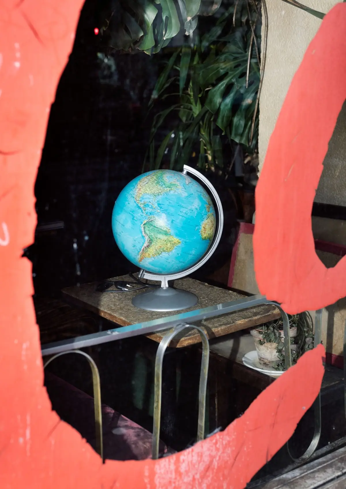 Blue world globe on a metal stand atop a wooden desk, framed by red torn-paper panels, with green tropical plants in the background.