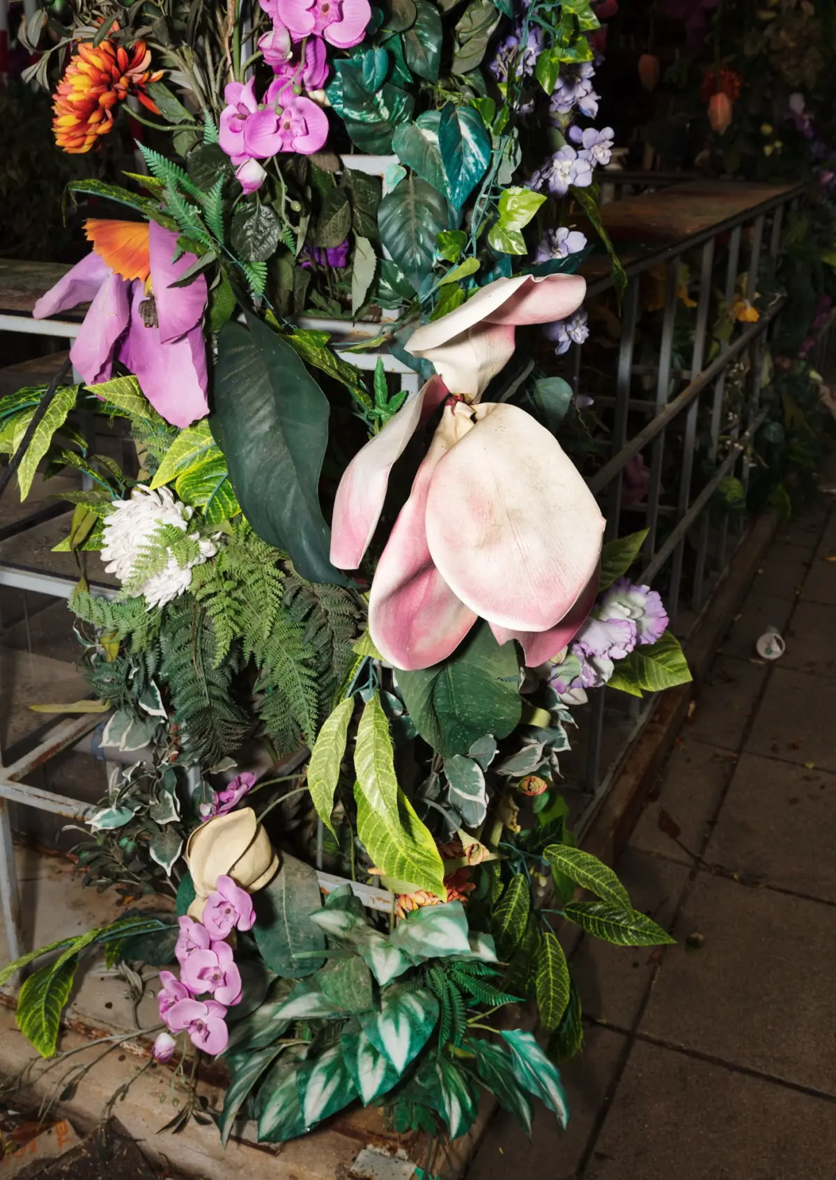 Artificial flower arrangement wrapped around a metal railing along outdoor stairs, featuring pink orchids, pink calla lilies, and green foliage.