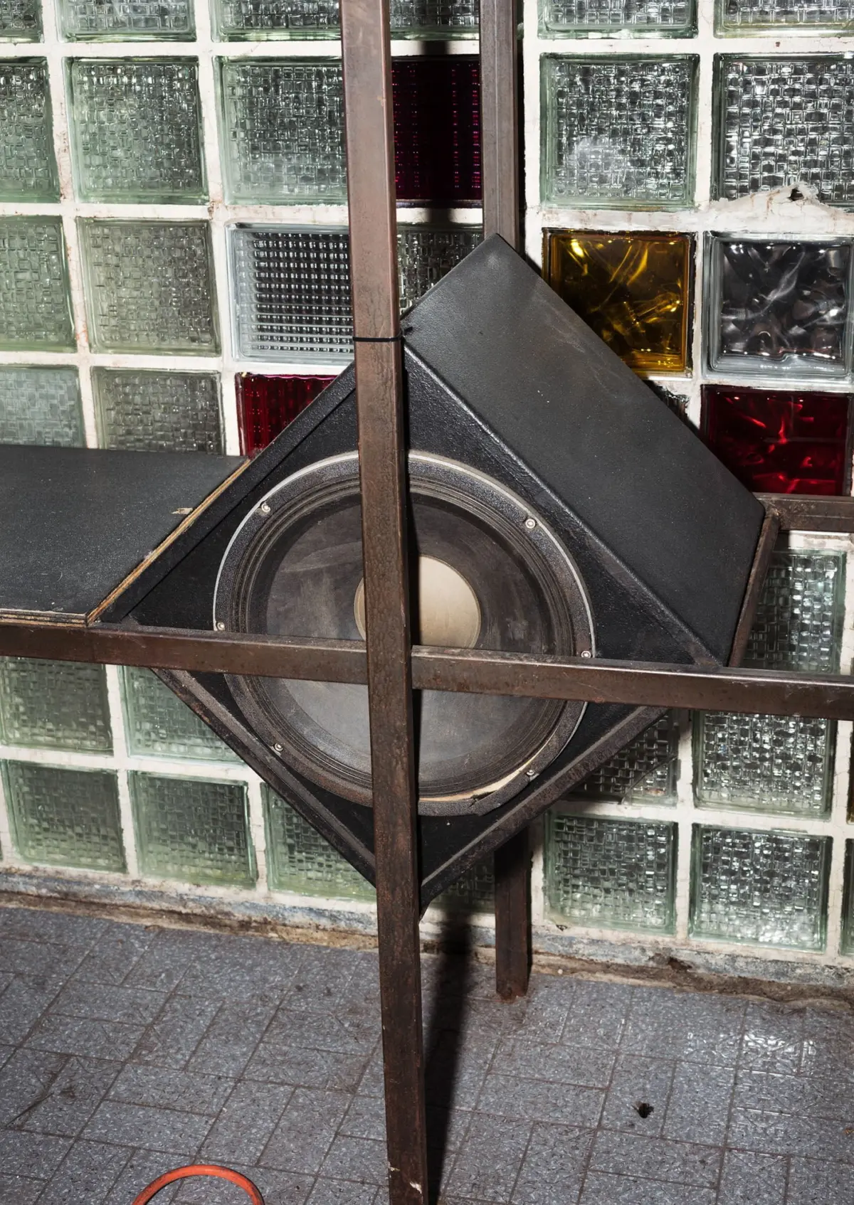 Diamond-shaped black subwoofer mounted in a wooden frame against a glass-block wall; orange cable on tiled floor.