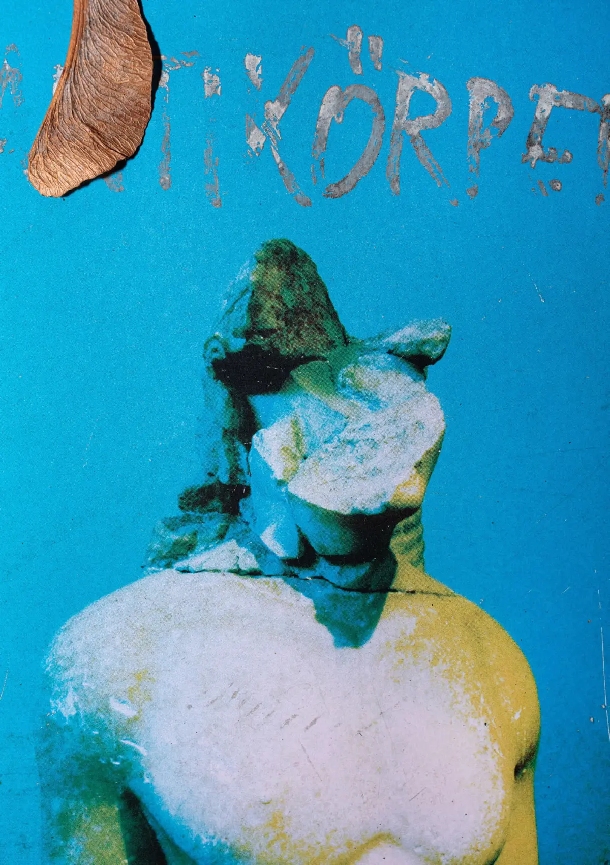 Weathered stone bust with green patina on head and neck against a bright blue background; a dried leaf rests to the left.