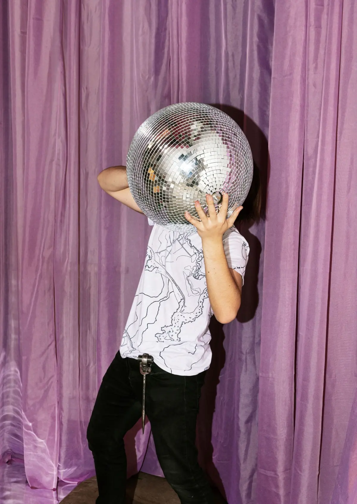 Person holding a large disco ball in front of their face, standing before purple velvet curtains.