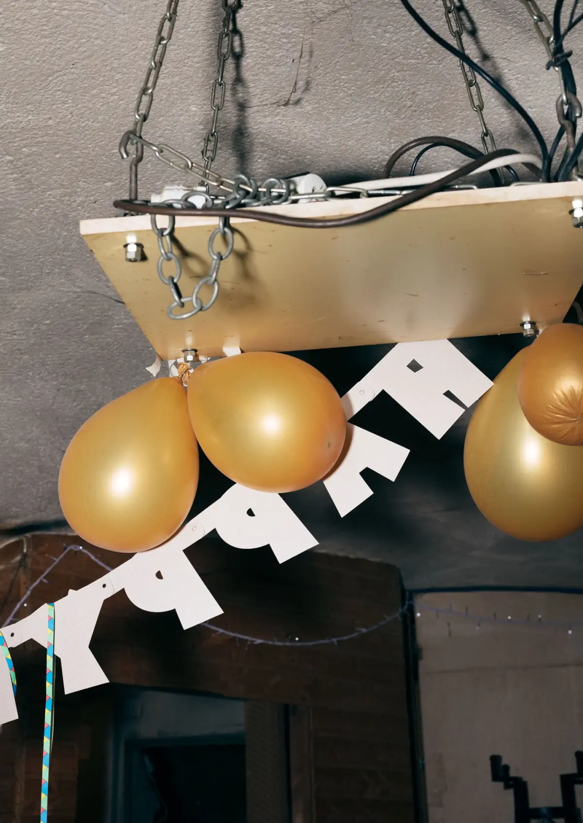 Gold balloons hang from the ceiling beside a white PARTY banner in an indoor party setup with exposed chains and wires.