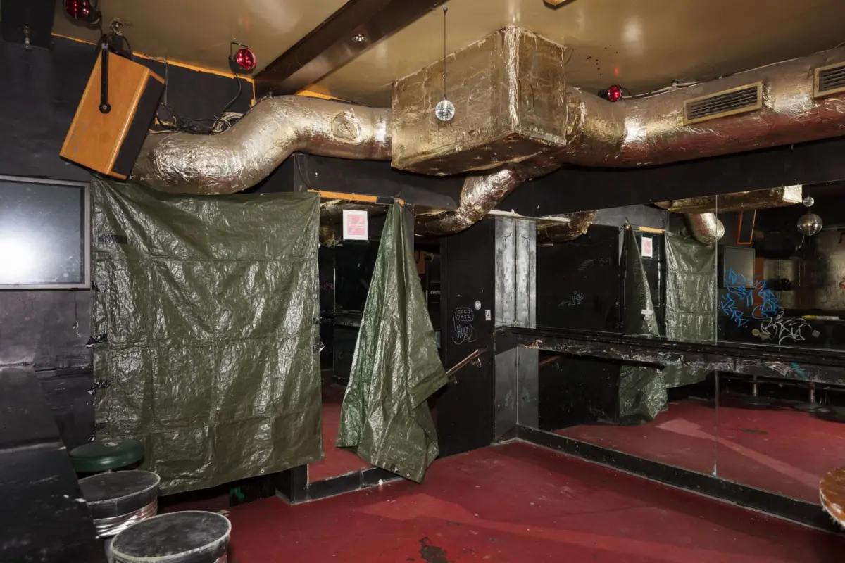 Empty nightclub interior with foil-wrapped HVAC ducts, hanging disco ball, graffiti walls, and green tarps as partitions.