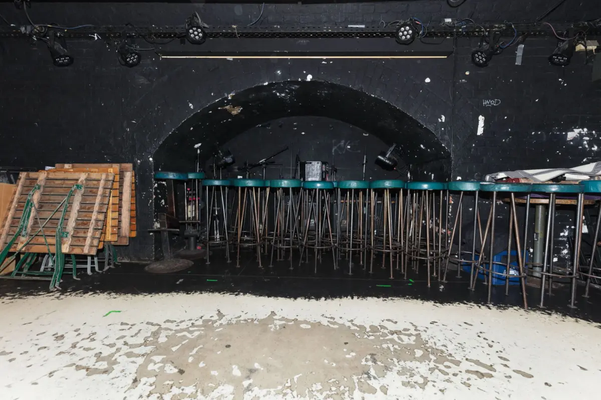 Backstage theater scene with folding chairs stacked under an arch, and wooden ladders on the left; paint-splattered floor.