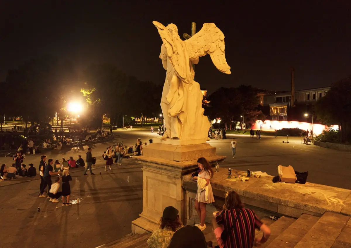 Stone angel statue with outstretched wings on a pedestal in a nighttime city square, with crowds gathered around.