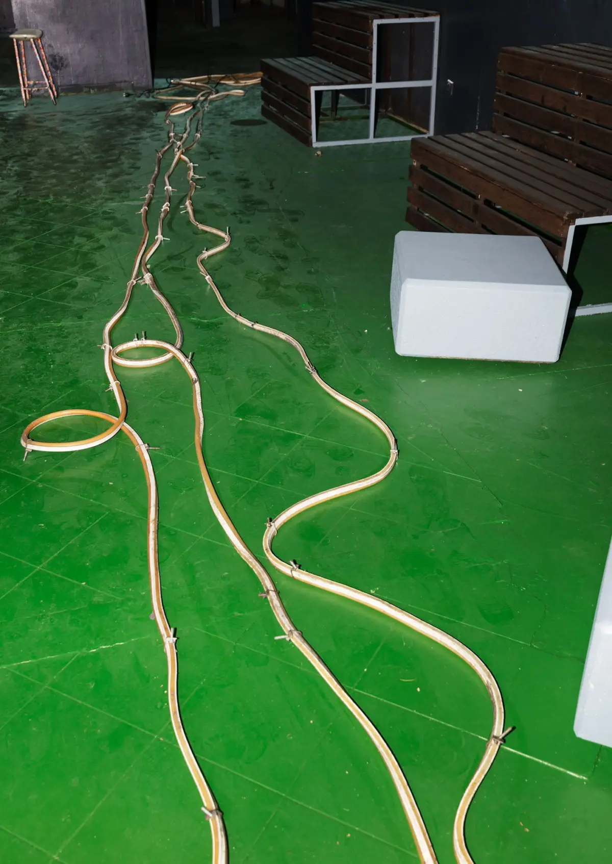 Long beige electrical cables snake across a bright green floor in a backstage studio, with wooden benches and a gray block seat in the background.