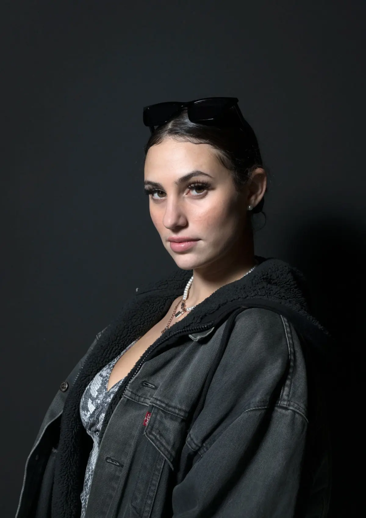 Portrait of a person with dark hair wearing a shearling-lined denim jacket, sunglasses atop head, pearl necklace, against a dark studio background.