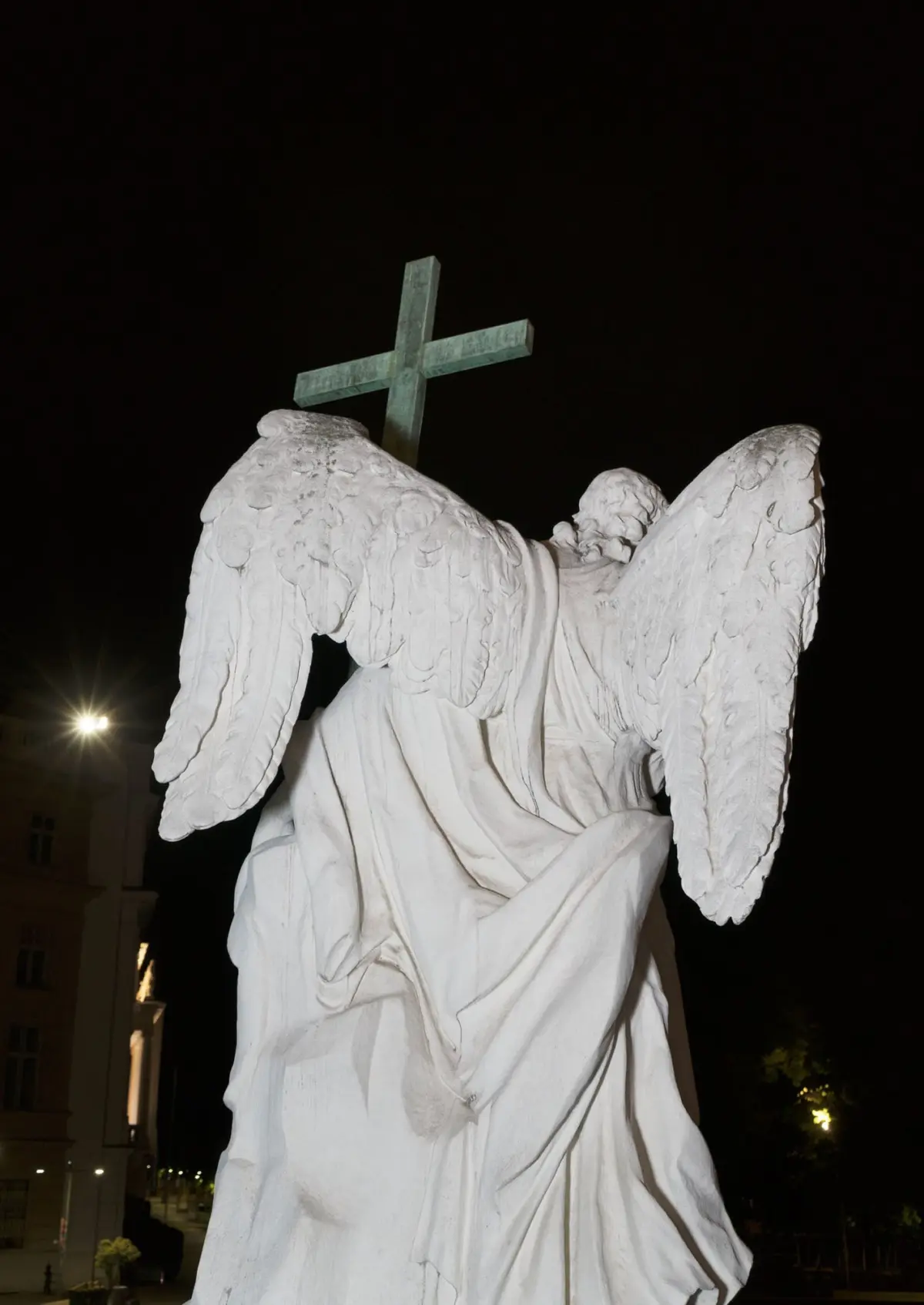 White angel statue with wings and a green cross atop a rocky pedestal at night.