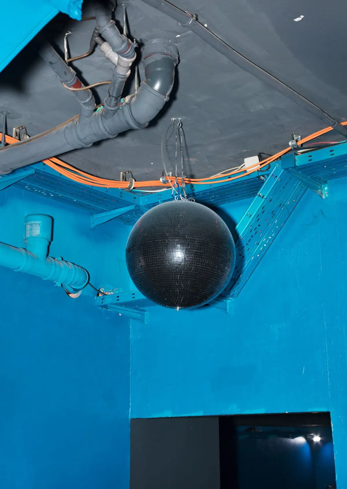Black disco ball hanging from the ceiling in a blue industrial room with exposed pipes and orange cables.