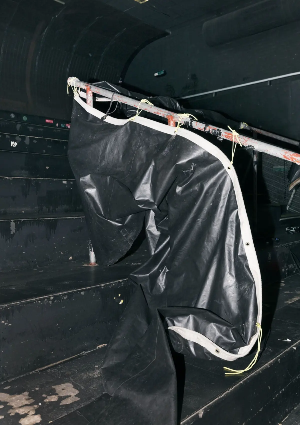 Black tarpaulin draped over a rusted metal frame, secured with yellow cords, in a dark curved industrial tunnel.