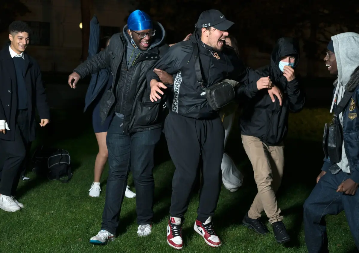 Group of young men dancing and laughing on a grassy lawn at night, wearing casual jackets and sneakers.