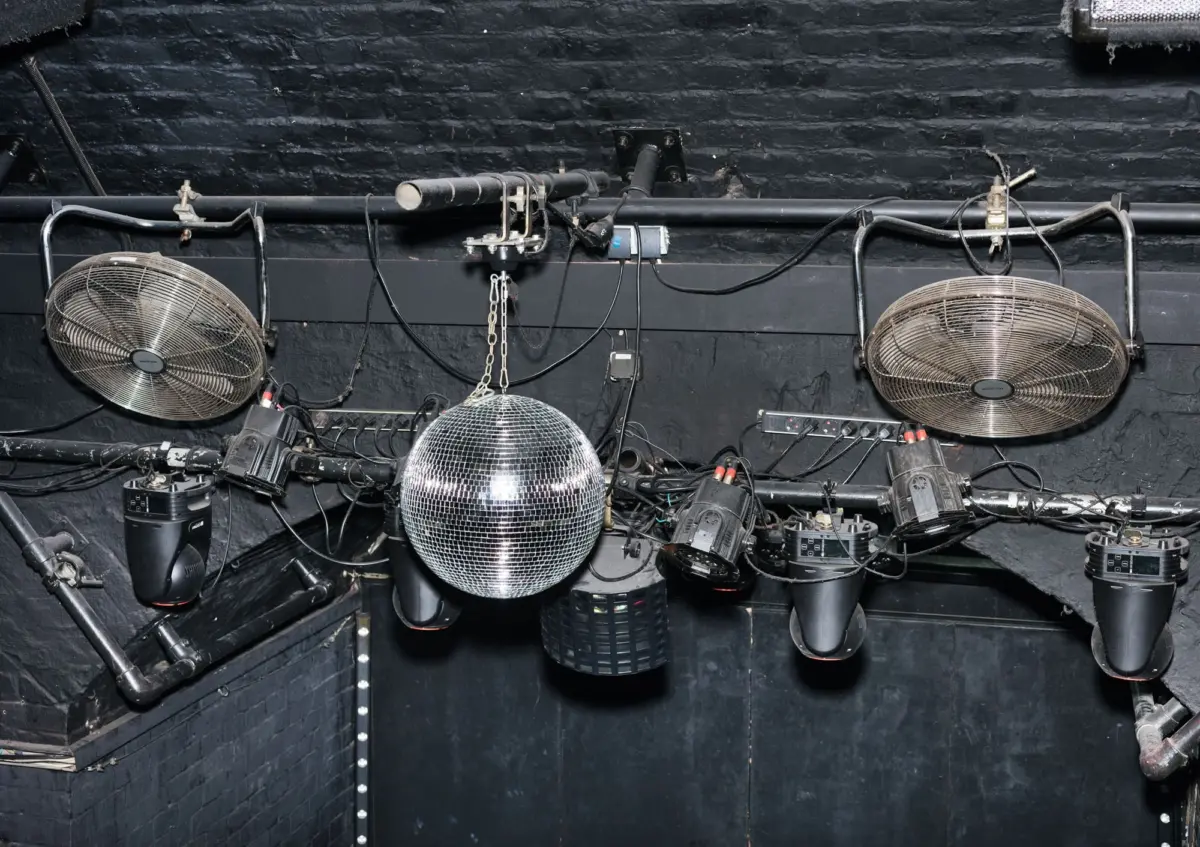 Disco ball suspended between two industrial ceiling fans on a black backstage wall, with tangled cables and lighting gear.