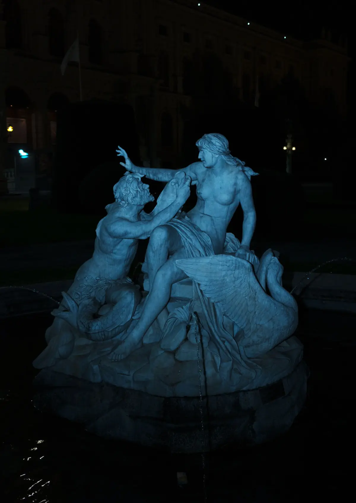 Blue-lit marble fountain sculpture: a muscular male grapples a winged female figure at night before a historic building.