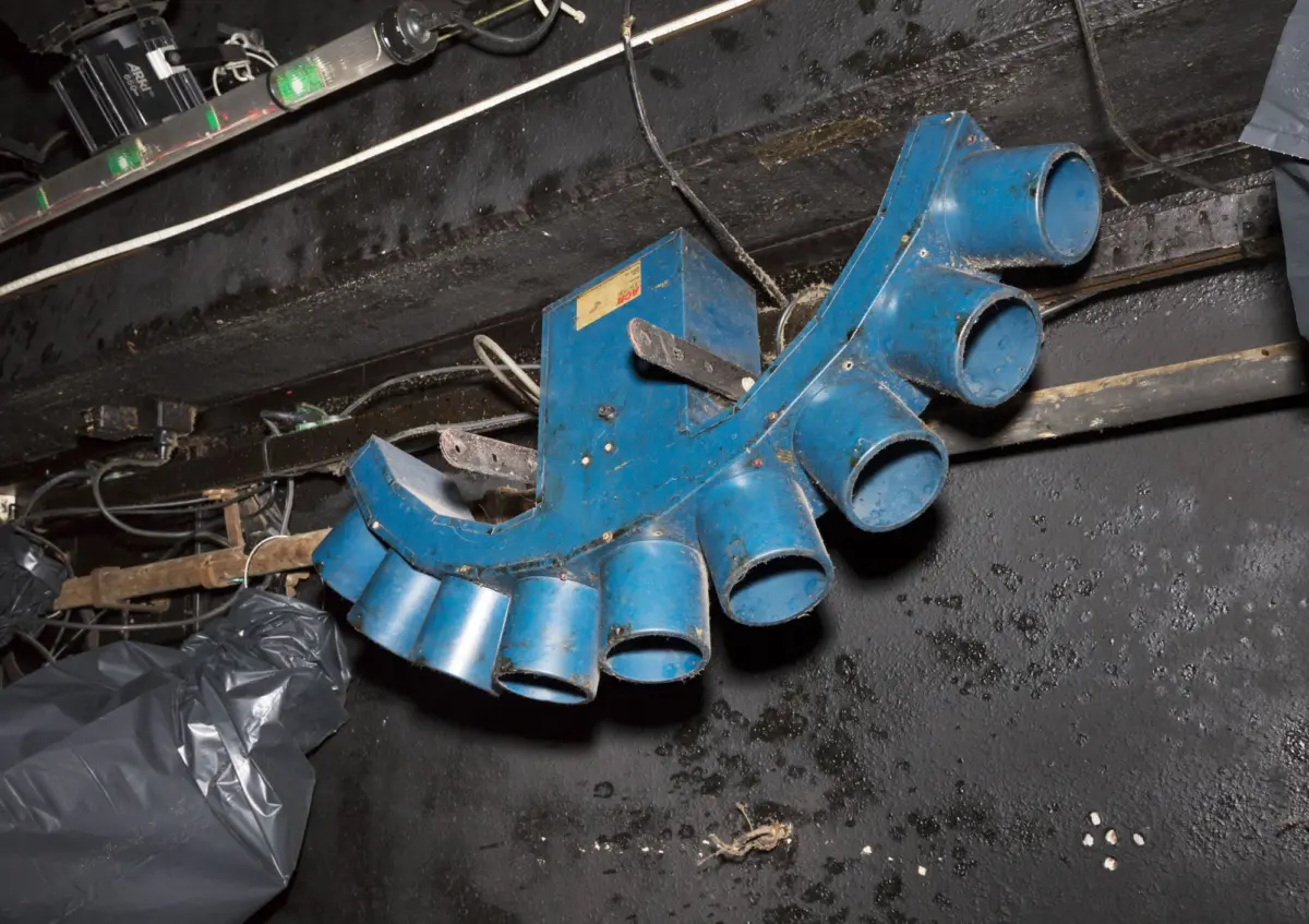 Blue metal ventilation manifold with four cylindrical outlets, resting in a dusty, cluttered industrial space.