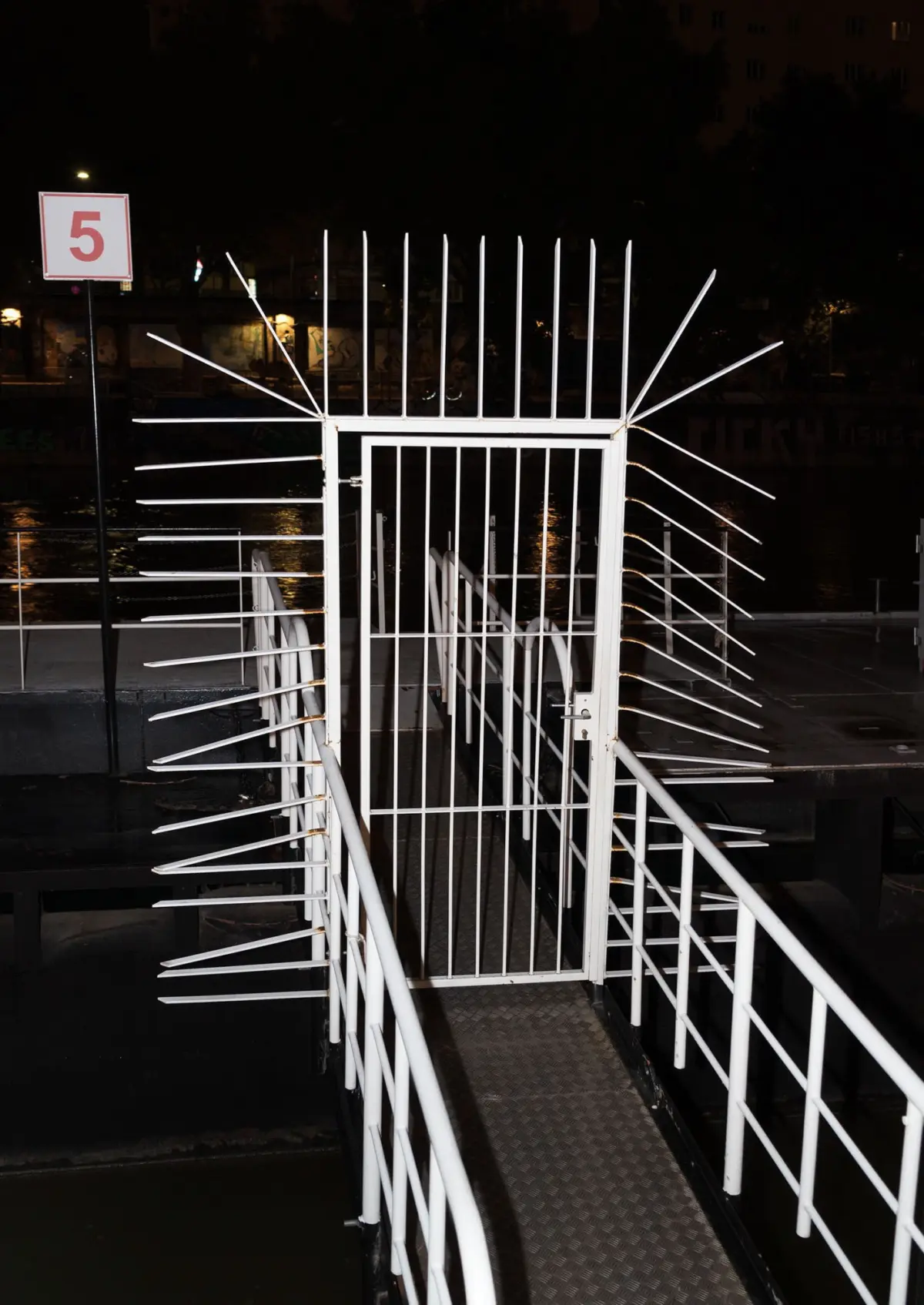 Nighttime view of a white metal gate with long outward-pointing spikes along a waterside walkway.