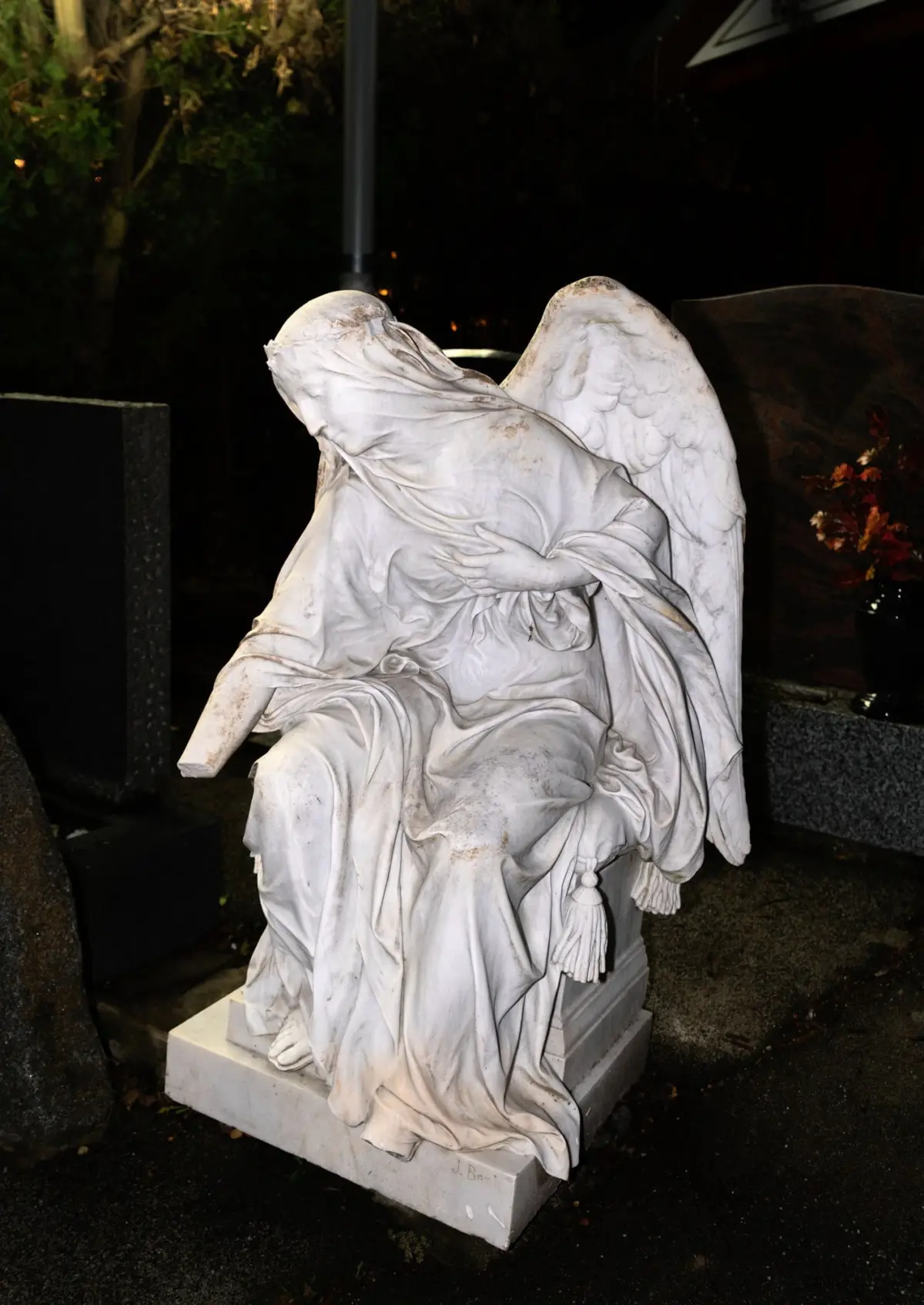 Seated marble angel statue with wings and flowing robes, head bowed, on a stone pedestal in a cemetery at night.