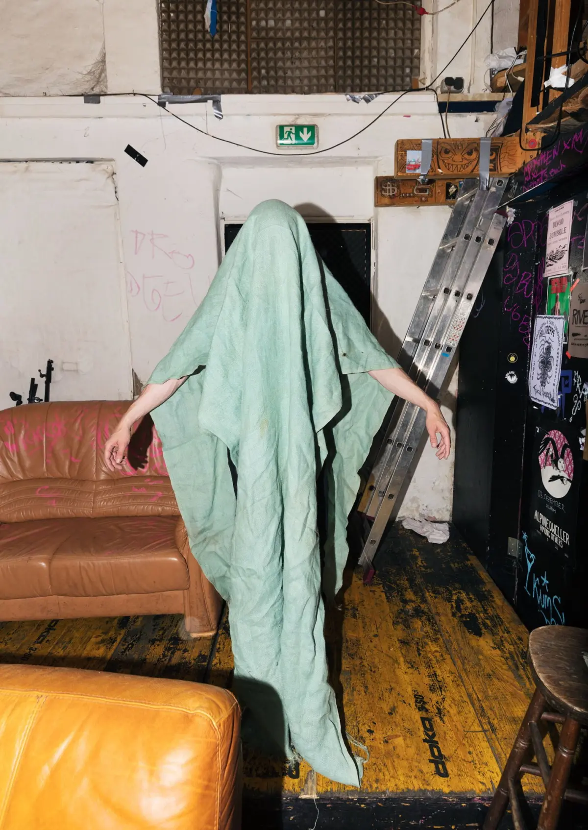 Person draped in a light-green sheet stands in a gritty backstage room with ladder, graffiti, and posters.