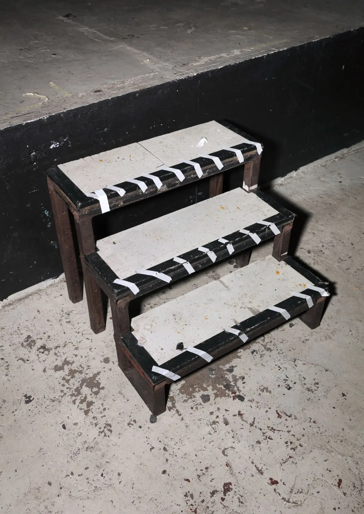 Three-step wooden staircase with white edge tape on each tread, set on a dusty concrete floor beside a black wall.