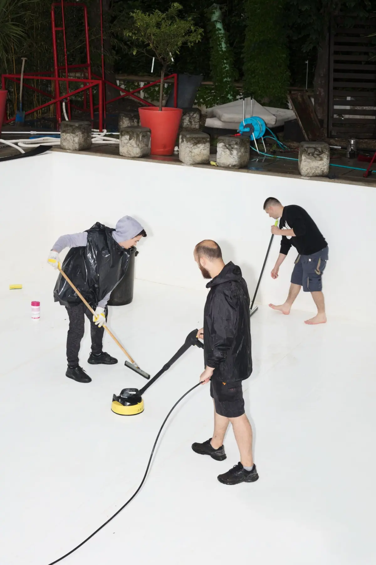 Three workers in black ponchos scrub an empty swimming pool with a floor-scrubbing machine; hoses and garden gear lie nearby.