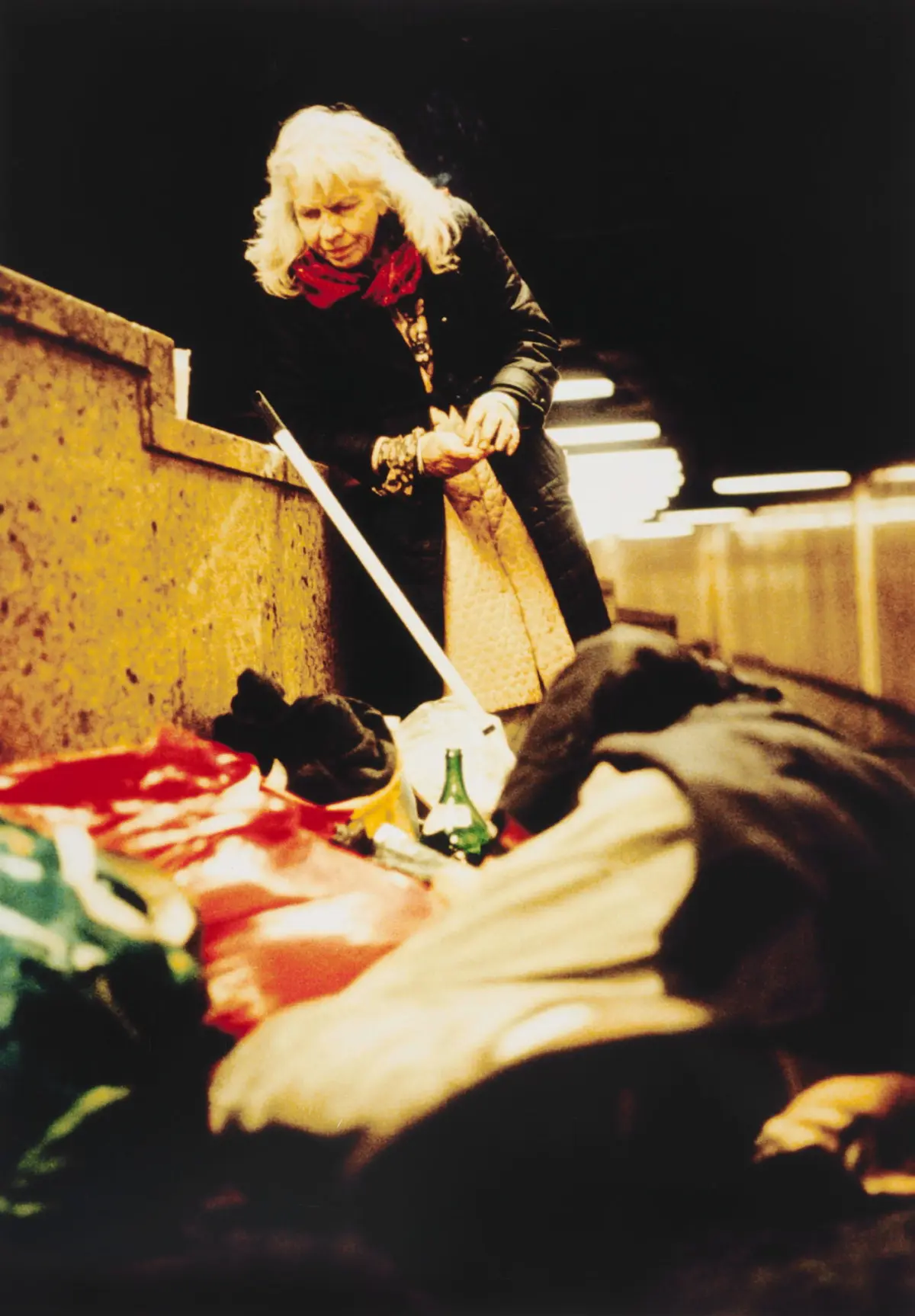 Older woman with long white hair and red scarf leans over a concrete barrier, rummaging through bags on a dimly lit street or platform.