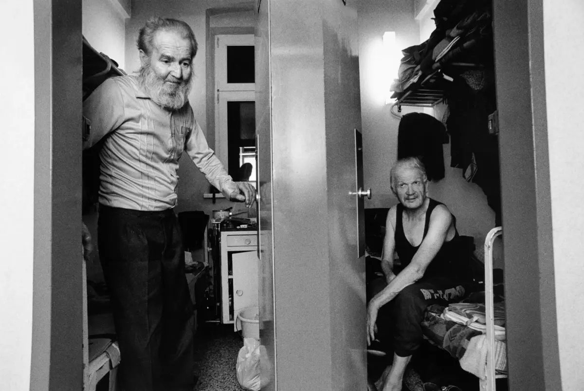 Two elderly men in a small dressing area; one with a long beard stands by open closet doors, the other sits on a bed among clothes.