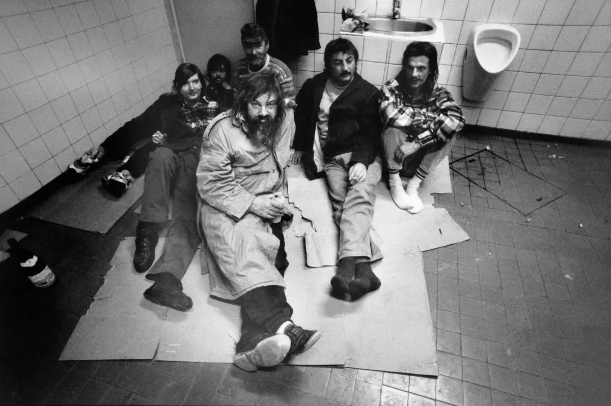 Group of six people with long hair and beards sitting on cardboard in a tiled bathroom; urinal visible on wall.