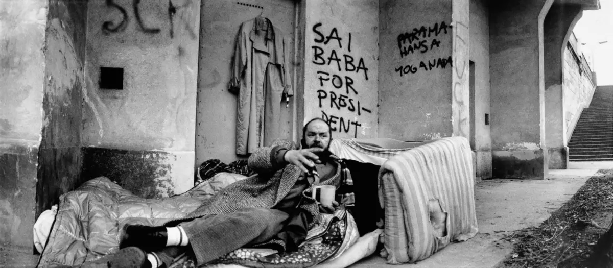 Homeless person lying on cushions and blankets on a sidewalk, reaching toward the camera; wall behind with graffiti SAI BABA FOR PRESIDENT; coat