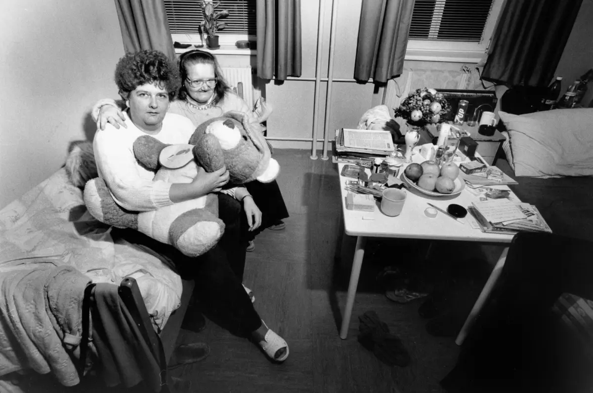 Two people sit on a sofa in a cluttered living room, hugging a large stuffed bear; table nearby holds fruit, magazines, and bottles.
