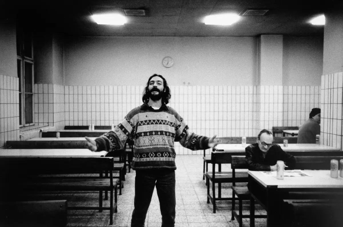 Bearded man in a geometric-patterned sweater stands with arms outstretched in a tile-walled cafeteria; a seated man at a nearby table.
