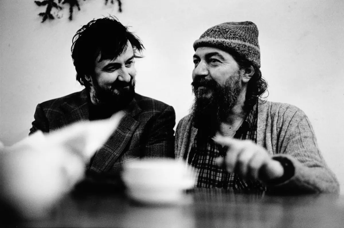 Two bearded men share a smile while seated at a table; dishes blur in the foreground.