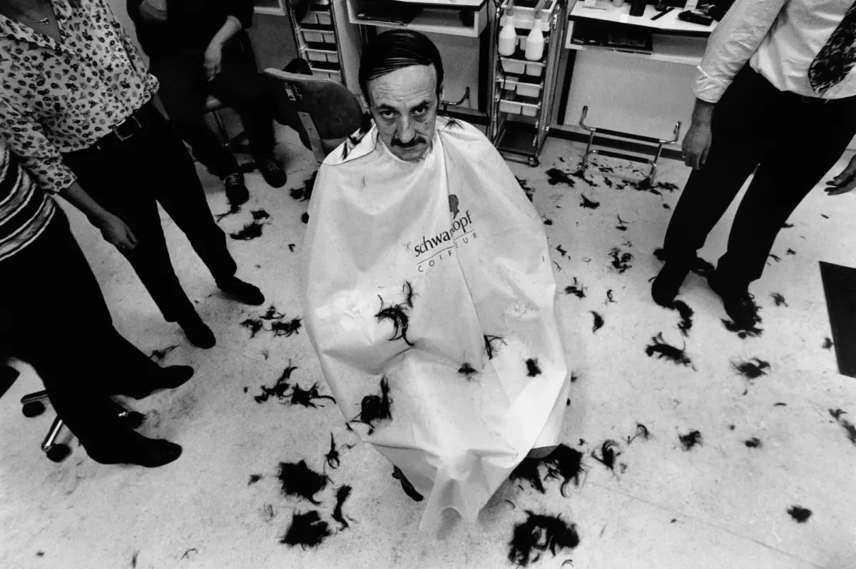 Man with slicked-back hair and mustache sits in barber chair wearing a Schwarzkopf cape; hair clippings on the floor.