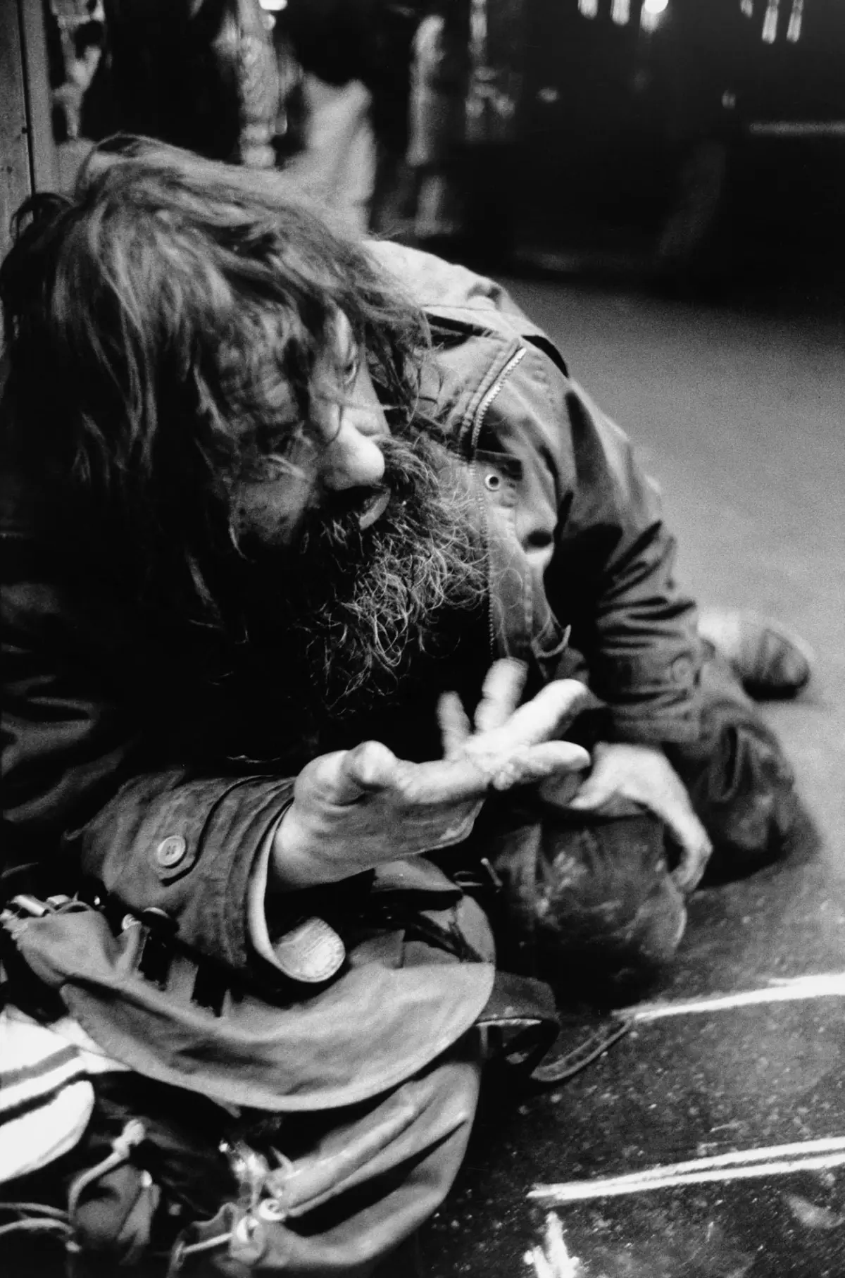 Adult person with long hair and beard, wearing a jacket, sitting on sidewalk, handling a rope.