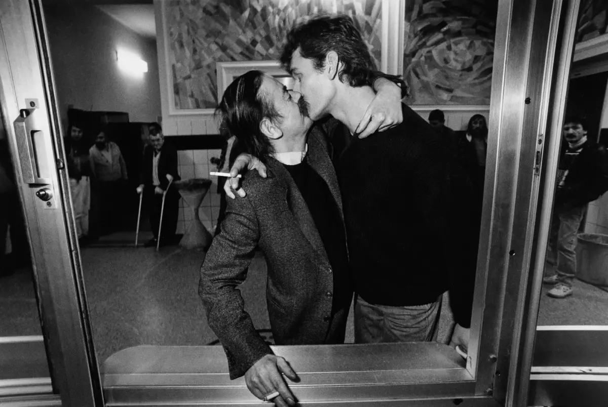 Two men kiss, one with arm around the other's neck, at a metal-framed doorway in a crowded room.