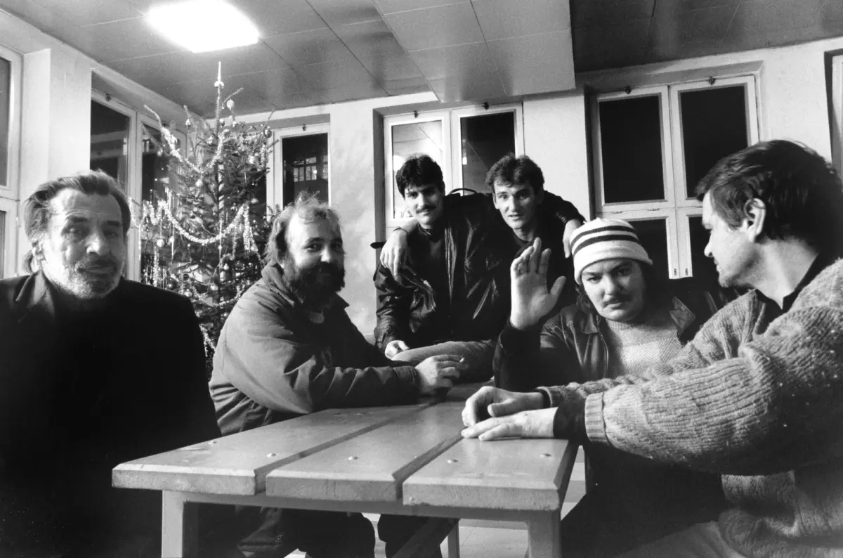 Group of adults around a wooden table in a room with a Christmas tree, some lean in and one person raises a hand.