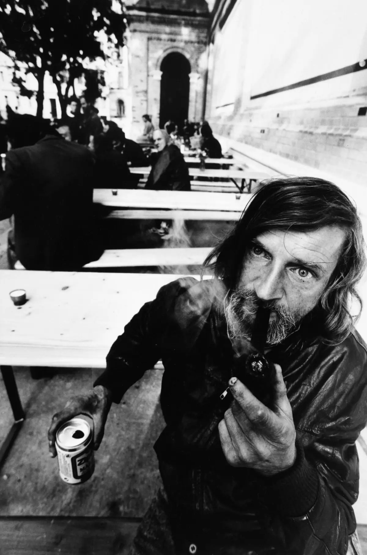 Bearded man with long hair smoking a cigarette at an outdoor cafe table; background shows other patrons and an arched entrance.