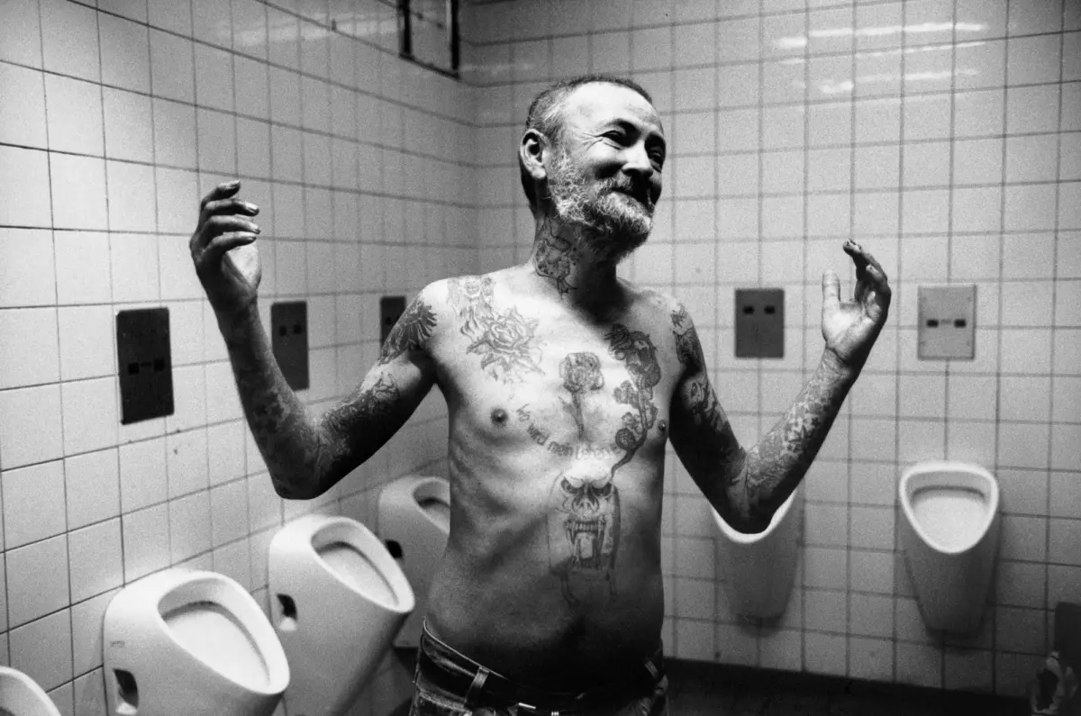 Shirtless adult with tattoos smiles, arms raised, in a tiled bathroom with urinals.
