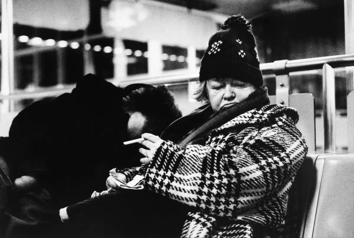 Person in knit beanie and plaid coat sits on a bus, looking down while holding a cigarette.