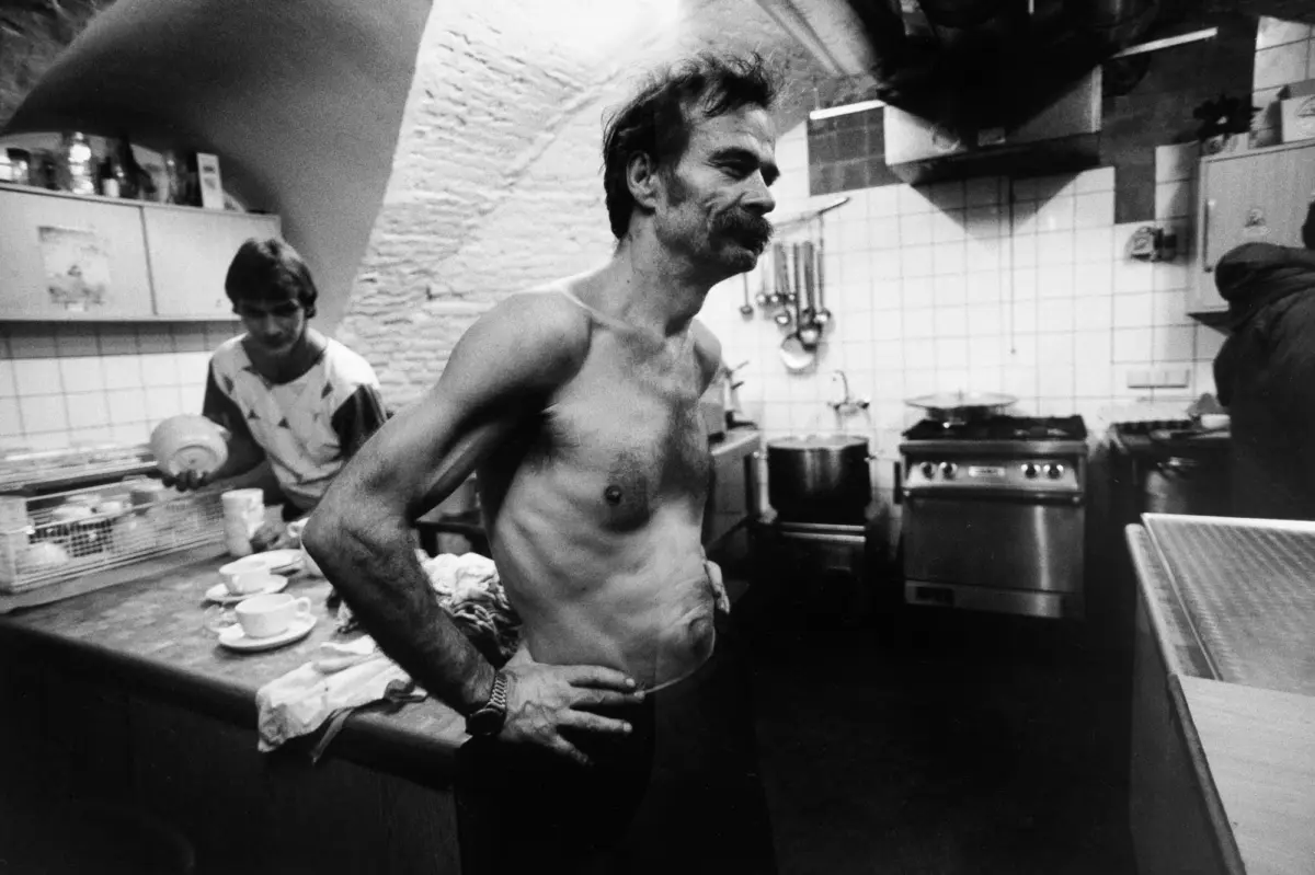 Shirtless adult with mustache stands with hands on hips in a busy kitchen; another person works at a counter amid ovens and hanging utensils.